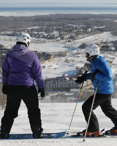 눈 덮인 언덕 꼭대기에 있는 두 사람이 블루 마운틴 리조트를 내려다보고 있습니다. 한 사람은 스노보드를 가지고 있고 다른 한 사람은 스키를 가지고 있습니다.