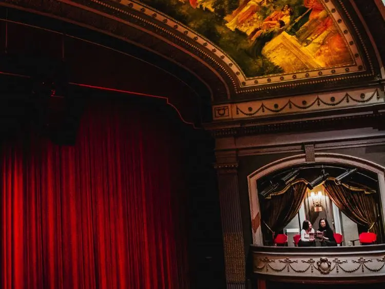 Dos mujeres en asientos del balcón lateral mirando hacia la cortina y el escenario en el Grand Theatre de Londres.