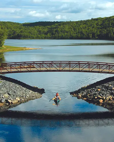 Two people in a canoe paddle under a foot bridge in Arrowhead Park.
