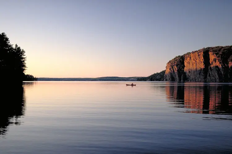 夕暮れ時の静かな湖を見下ろす崖と、一人のカヤック乗り。