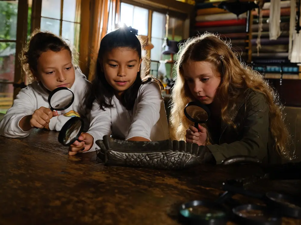 Three young girls examine an old item with magnifying tools.