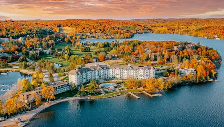  Vista aérea de Deerhurst Resort en Muskoka, enmarcada por las tranquilas aguas del lago y un vibrante bosque otoñal bajo un espectacular cielo rosa-naranja.