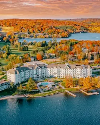 Vue aérienne du Deerhurst Resort à Muskoka, encadrée par les eaux calmes d’un lac et une forêt d’automne, sous un ciel rose-orange spectaculaire.