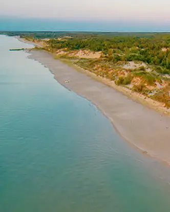 Luftaufnahme des Ipperwash Beach in Ontario in der Abenddämmerung, mit sanften Wellen, die an einen langen Küstenabschnitt mit Sanddünen und dichtem Wald im Hintergrund plätschern.