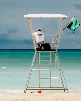 Ein Rettungsschwimmer sitzt auf einem hohen weißen Stuhl mit Blick auf das türkisfarbene Wasser von Grand Bend Beach, während eine grüne Flagge im Wind flattert.