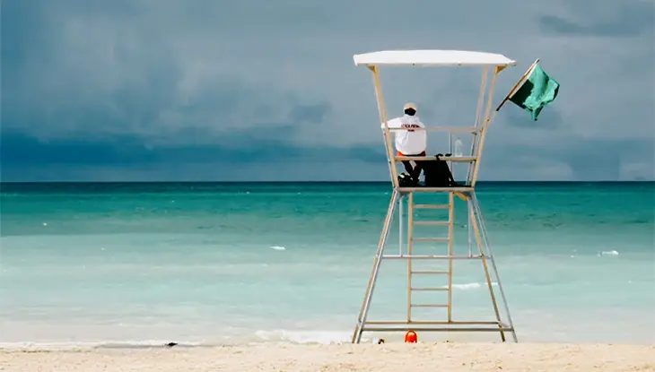 푸른색 바닷물이 펼쳐지는 그랜드 벤드 해변을 바라보며 키가 큰 흰색 의자에 앉아 있는 구조원. 바람에 녹색 깃발이 펄럭입니다.