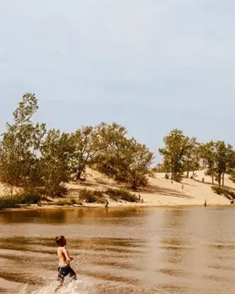  うねる砂丘、点在する木々、そしてかすんだ夏の空を背景に、サンドバンクス州立公園で水しぶきを上げる子供。