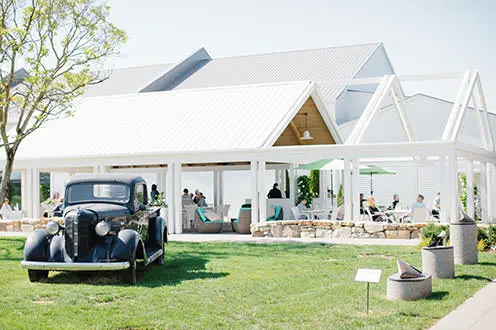Voiture noire d'époque stationnée sur l'herbe devant un grand gazebo blanc en plein air, où des gens sont assis sur des chaises et boivent du vin.