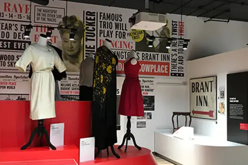 Joseph Brant 박물관의 실내 의류 전시. 방 안은 다양한 드레스로 꾸민 마네킹들로 가득하다. 여성의 사진이 전시회의 인용문과 제목과 함께 뒷벽을 따라 늘어서 있습니다.