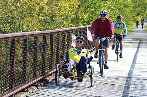 Cyclists crossing over a bridge on the Trans Canada Trail on a sunny day. There are pedestrians walking on the gravel trail in the background surrounded by lush greenery.