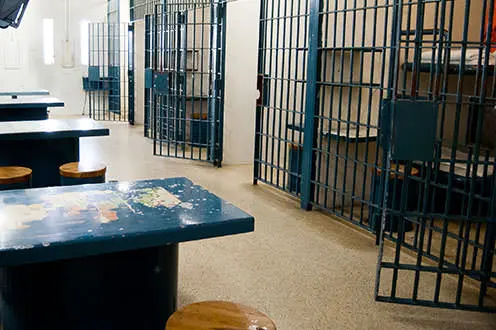 역사적인 SDG 감옥 내부의 열린 감방들이 늘어선 복도. 왼쪽에는 파란색 금속 테이블과 의자들이 놓여 있습니다.