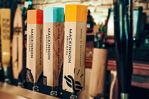 Tres grifos de cerveza de madera etiquetados con diferentes variedades de cerveza Mackinnon Brothers. Crosscut Canadian Ale, Origin Anceint Grain Ale y Bound By Brotherhood con etiqueta roja, azul o amarilla