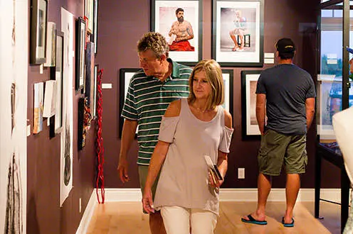 A man and a woman looking at the art displayed on the wall of an art gallery. Another man is shown in the background looking at art.