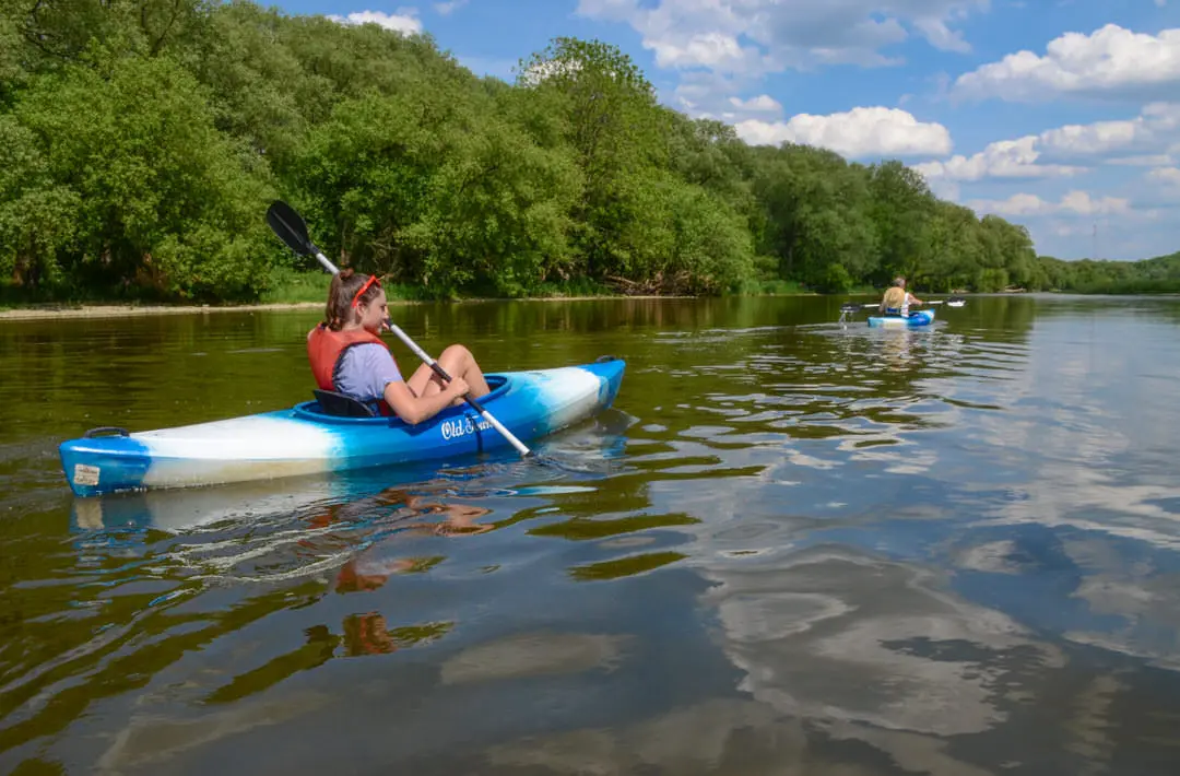 Femme pagayant en kayak sur une rivière bordée d'arbres et qui suit un second kayak.