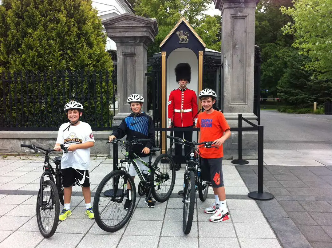 Trois jeunes enfants qui sourient et qui marchent à côté de leur vélo à côté d'une auto-patrouille de la GRC stationnée entre deux colonnes de pierre.
