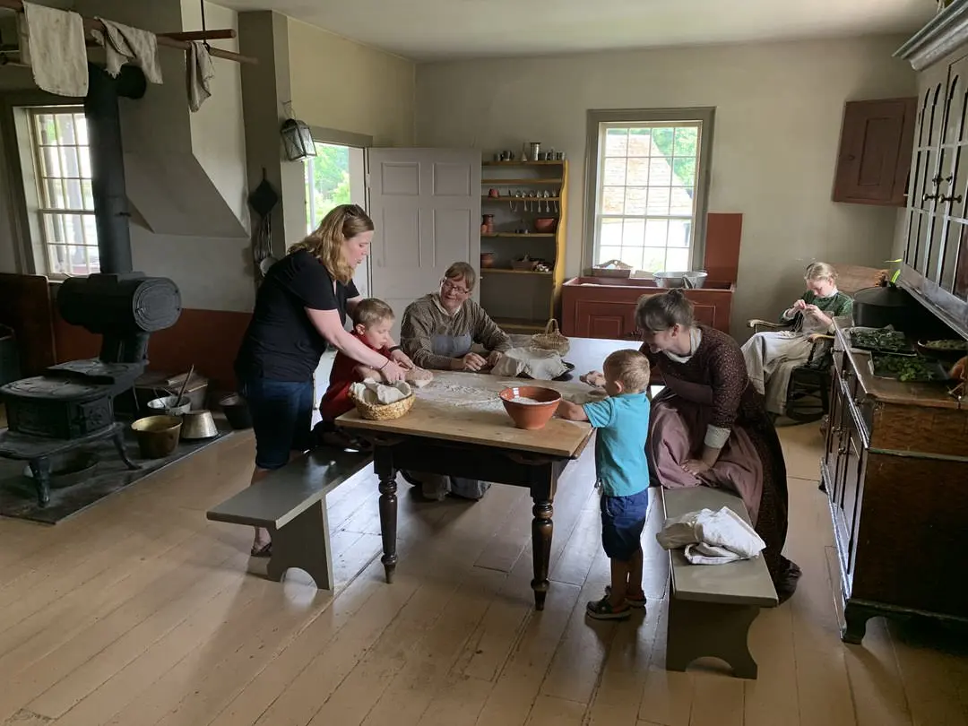 3 mujeres con vestidos largos de pioneras con 2 de ellas en la mesa de la cocina ayudando a una mujer moderna y 2 niños pequeños a amasar pan