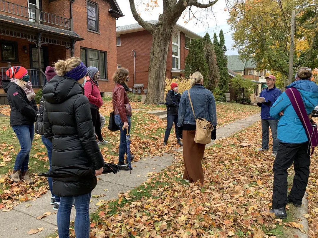 Huit personnes debout sur le terrain d'une ancienne maison sur un tapis de feuilles d'automne et qui regardent un guide touristique qui s'adresse à eux.