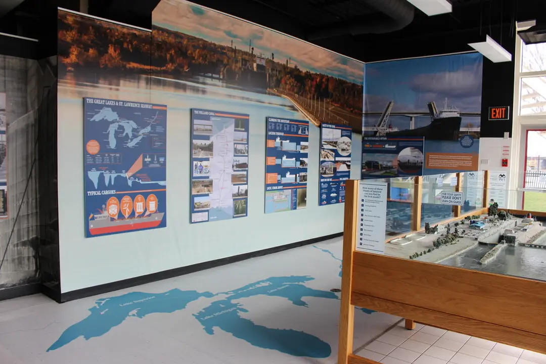 À gauche, écran mural de cartes et de photos, et à droite, grande photo d'un vaisseau dans une écluse au-dessus d'un modèle réduit d'une écluse.