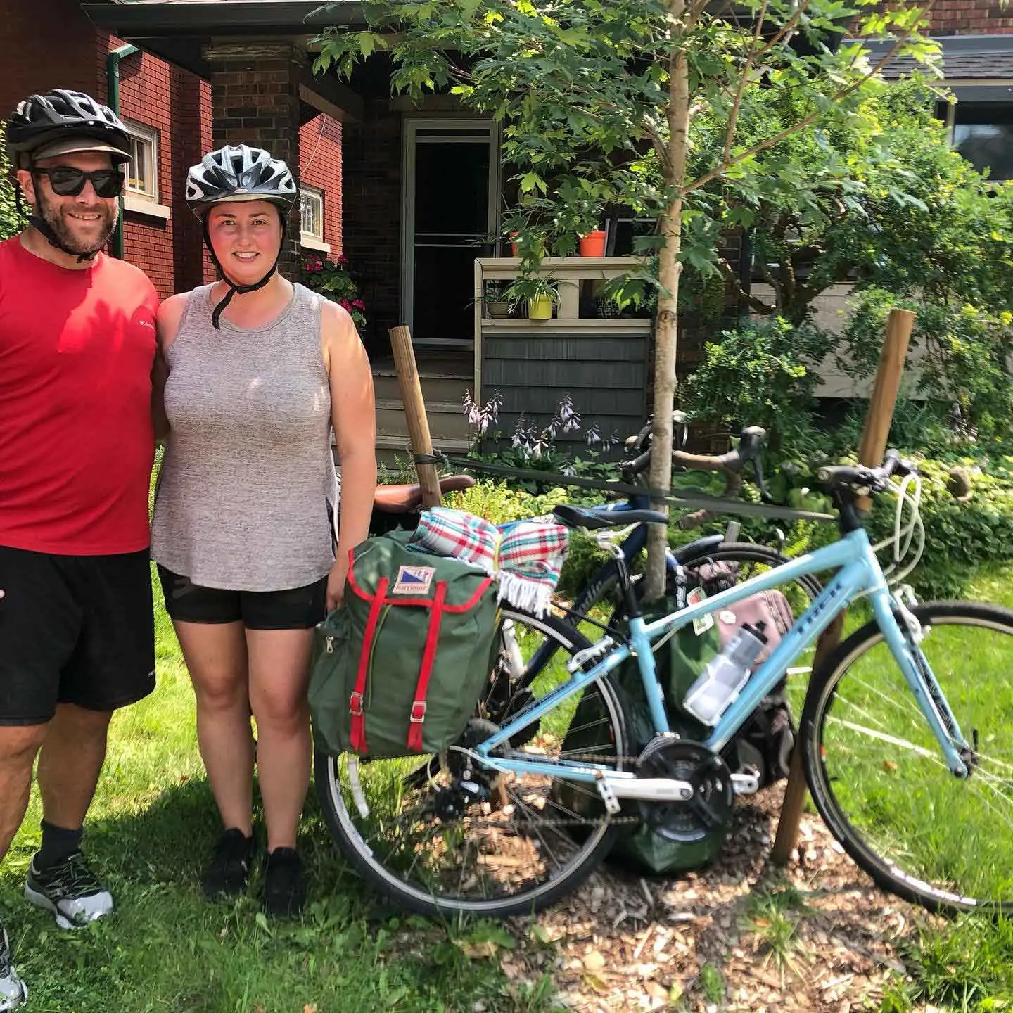 A man and a woman, both wearing bike helmets, pose with a bike that is leaning up against a tree