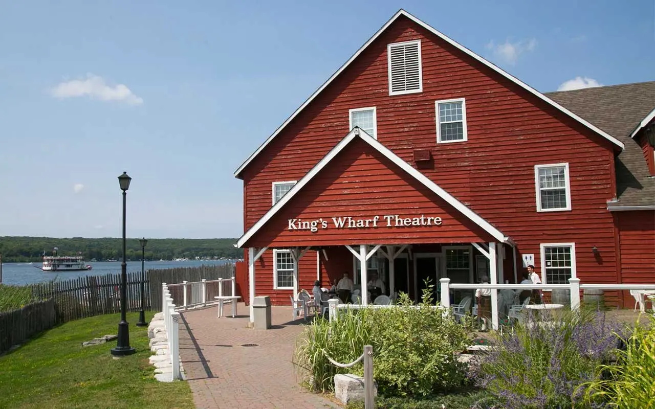 Personnes qui discutent à l’extérieur d’un bâtiment en bois rouge situé au bord de l’eau.