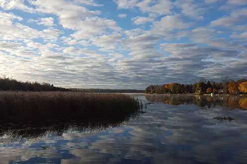 A scenic view of the lake on a cloudy day with the shore in the distance. There is a clear reflection of the sky in the water. 