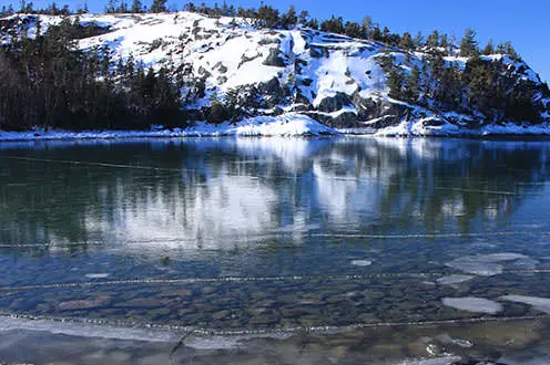 A scenic view of a partially frozen lake with a rocky snow covered hill in the distance.
