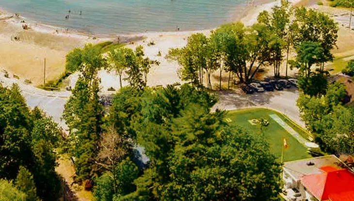 Vista aérea de Balm Beach Resort, con visitantes disfrutando de la costa arenosa y aguas poco profundas, rodeados de frondosos árboles y pequeños edificios.