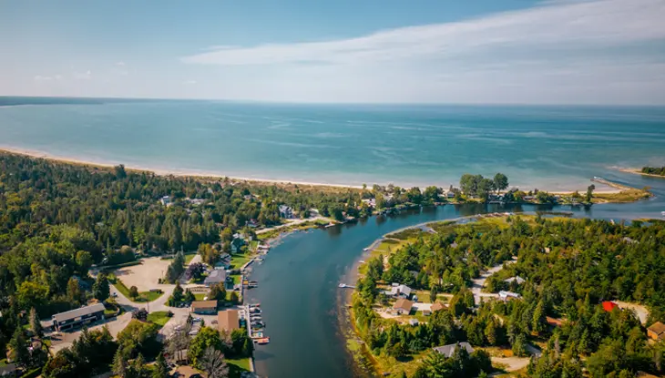 Luftaufnahme der Mündung des Sauble River in den Huronsee bei Sauble Beach, Ontario, mit von Bäumen gesäumten Cottages, sandigem Ufer und klarem blauen Wasser.