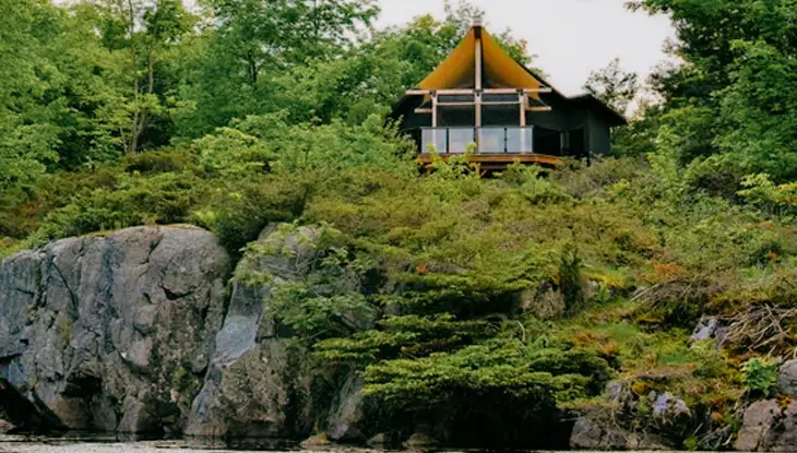 A-frame cabin in Pine Brae Wilderness Escape perched above the water on a rocky shoreline, surrounded by dense green forest.