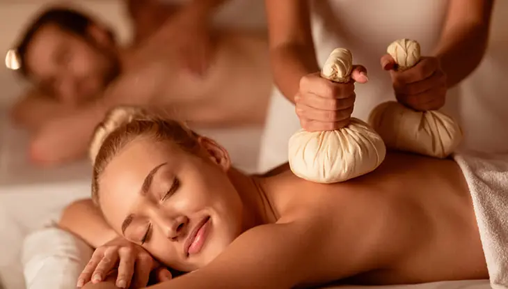 Femme satisfaite étendue sur une table de massage tandis qu'une massothérapeute appuie sur deux sacs de tissu blanc sur son dos. 