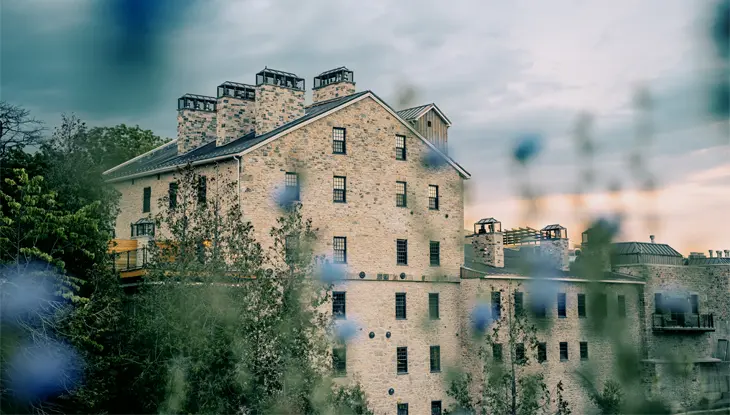 Das rustikale Elora Mill Hotel & Spa aus Stein mit mehreren Schornsteinen, zu sehen durch zartblaue Wildblumen unter einem dramatischen Abendhimmel.