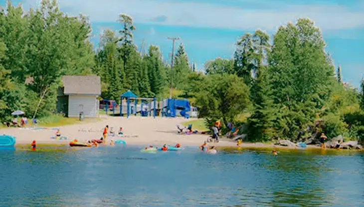 Familien genießen einen Sommertag an einem der Sandstrände von Kenora, während die Kinder in der Nähe der Küste schwimmen und spielen, umgeben von Wald.