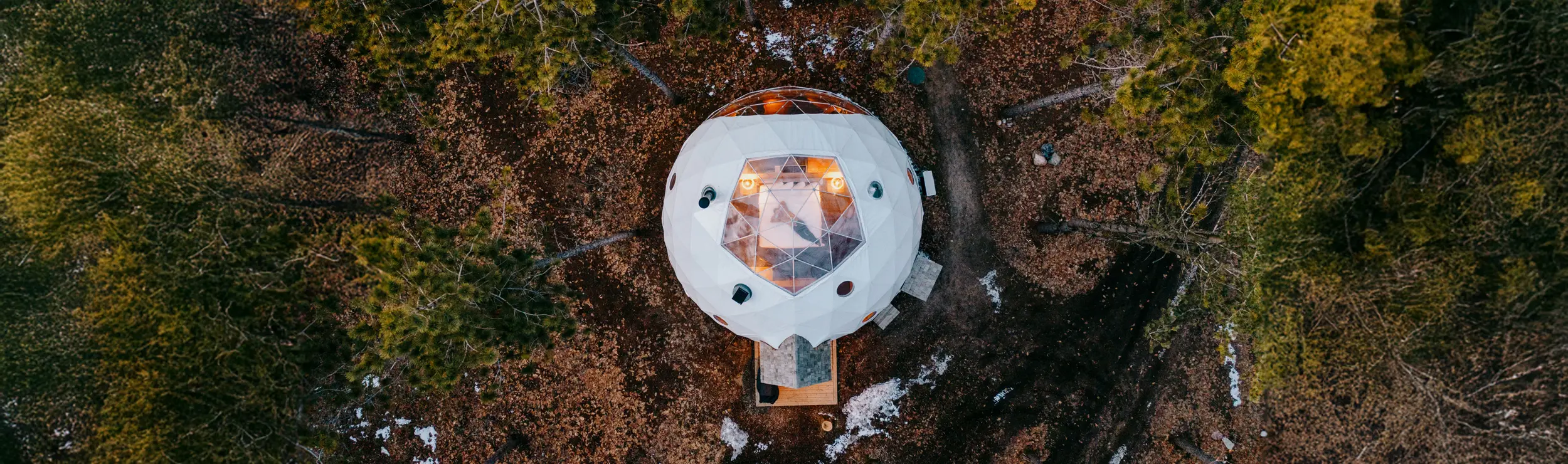 Geodesic dome nestled among trees in Glen Oro, glowing warmly in a serene, snow-dusted forest.