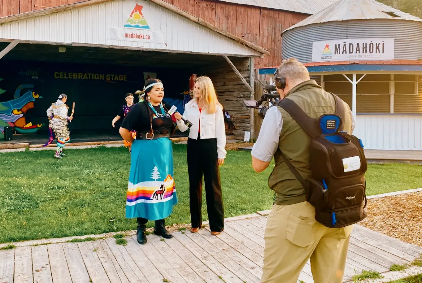 A person in traditional attire being interviewed at Mādahòkì's Celebration Stage on Mādahòkì Farm, Nepean, while dancers perform in the background.