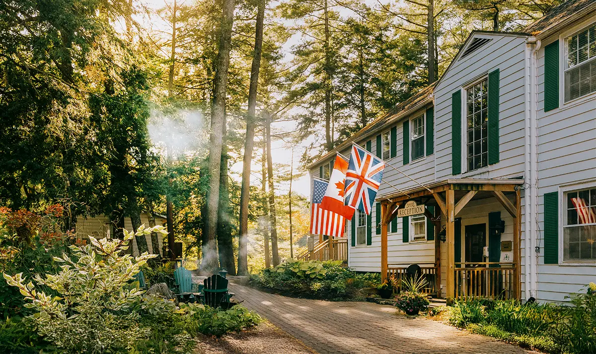 Sherwood Inn à Port Carling, avec de grands arbres, des chaises Adirondack vertes et des drapeaux internationaux, éclairé par la lumière du soleil.