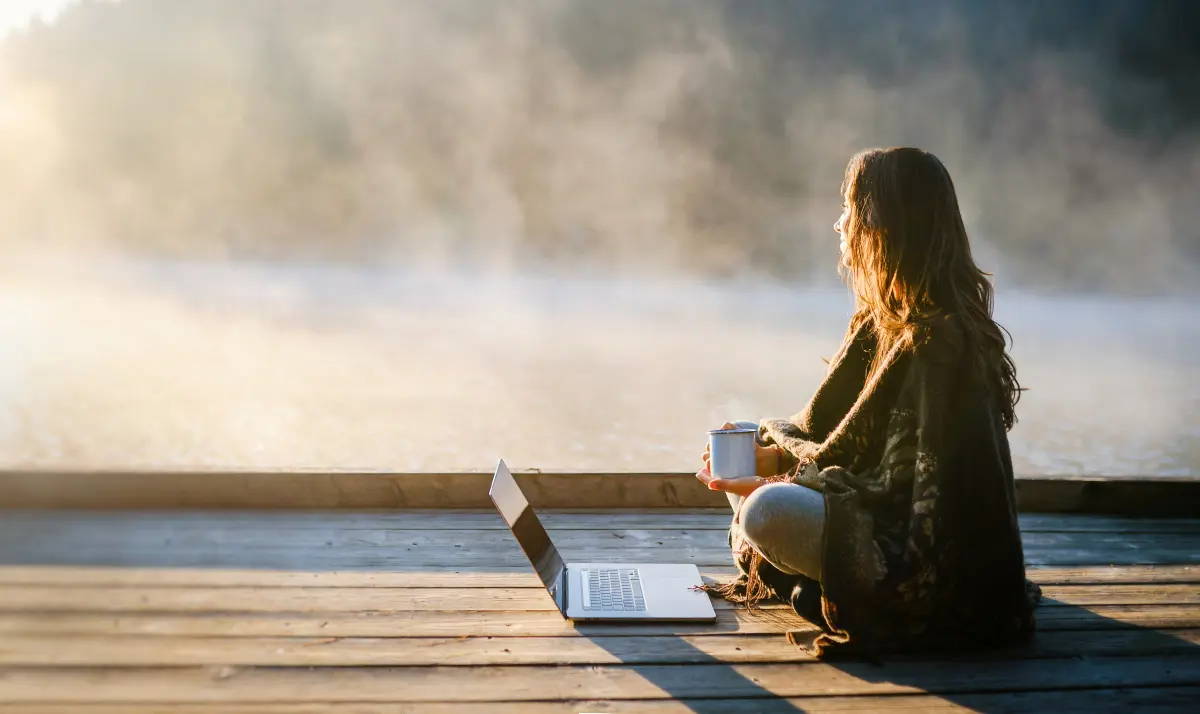 A young woman working on a laptop, sitting cross-legged near a misty lake and lush hills in the background. 