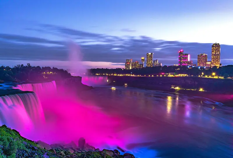 空が夕暮れに変わり、高いホテルの建物から光が輝く中、ピンク色の光が滝を照らします。