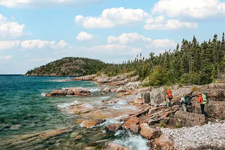Hikers walking along a rocky shoreline on Lake Superior with forested hills in the background.