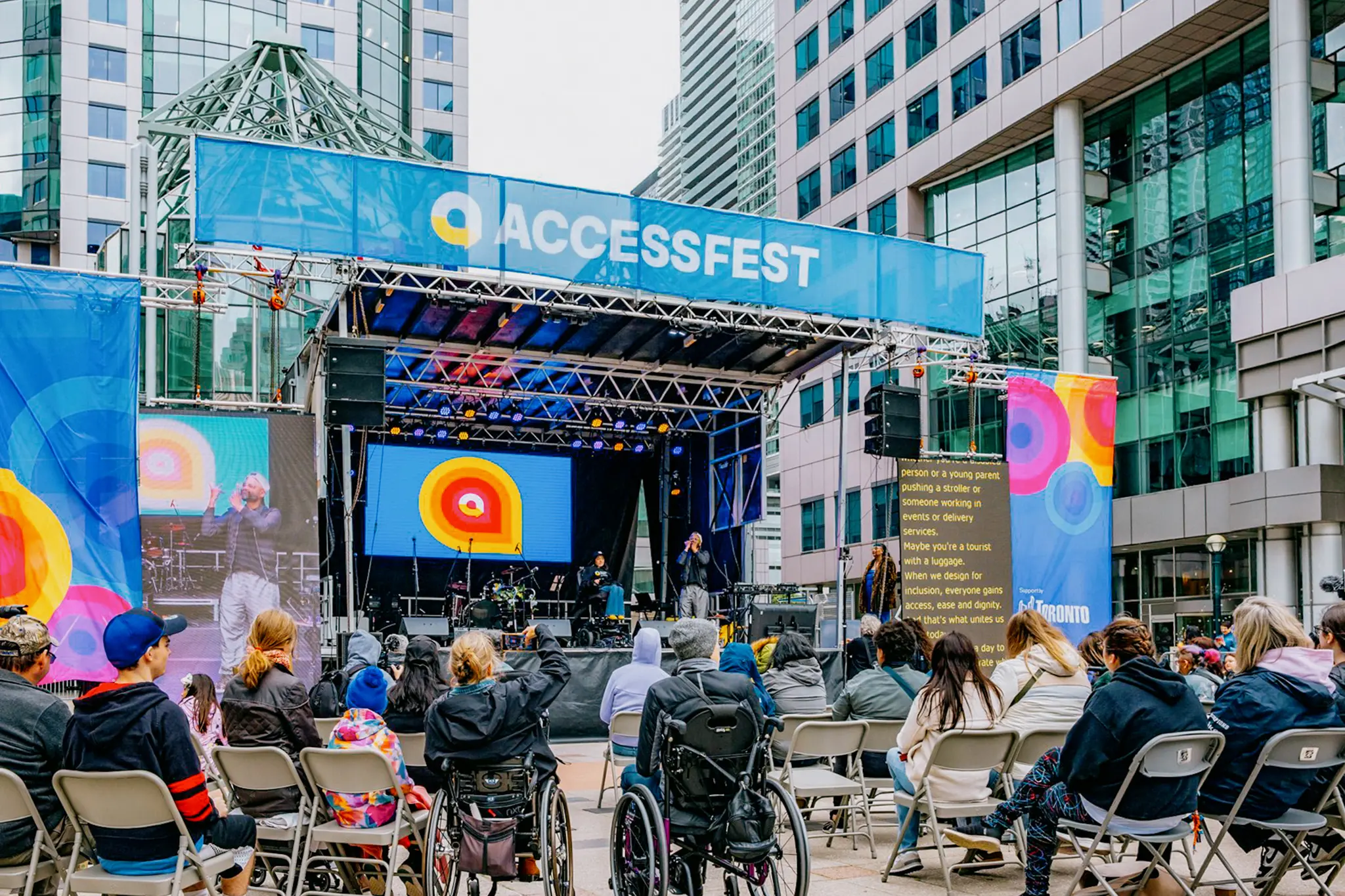 Une foule diversifiée, comprenant des personnes en fauteuil roulant, assiste à un spectacle en direct sur une scène extérieure à AccessFest, avec des tours de bureaux en verre en arrière-plan.