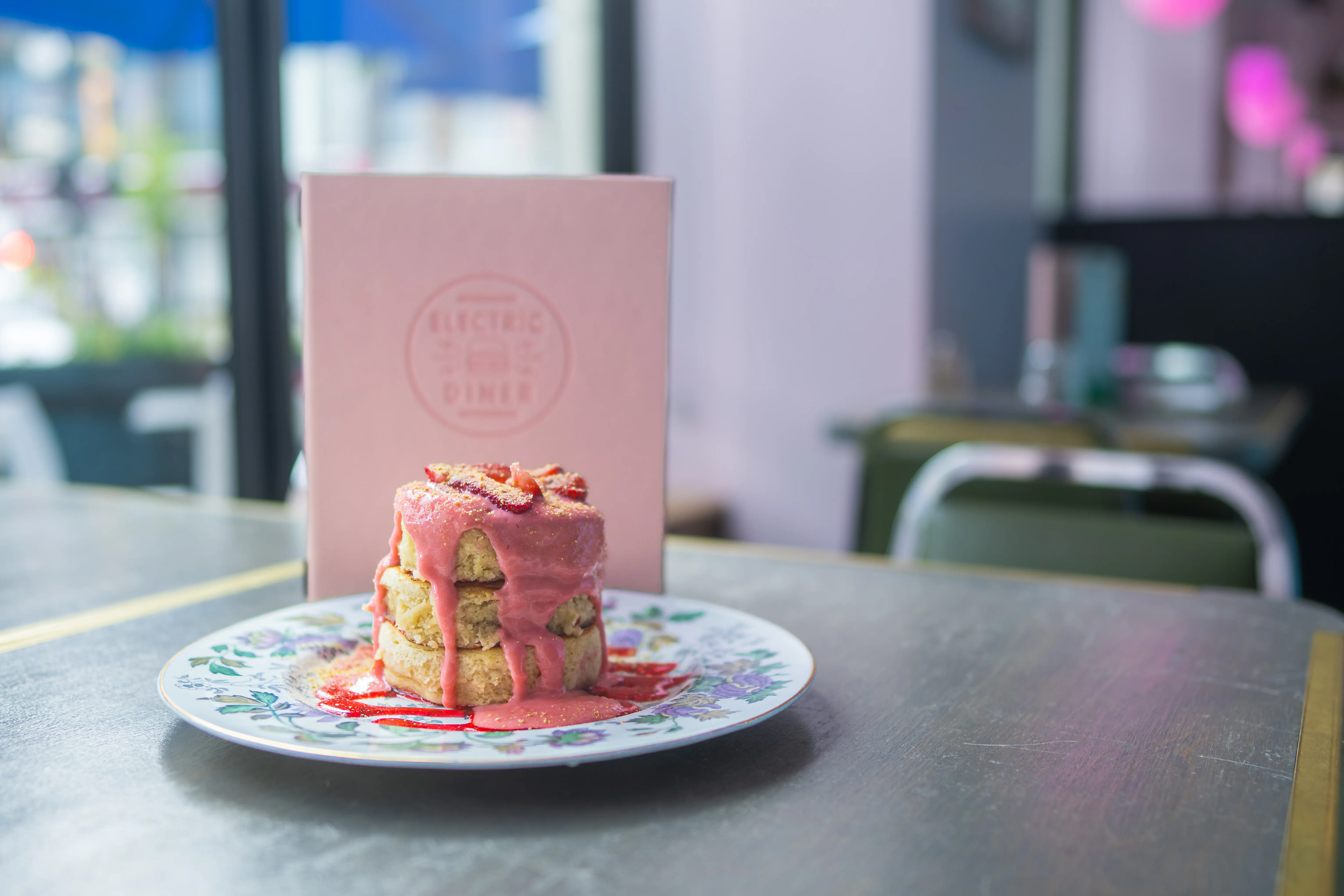 Tres grandes gofres con capas de salsa de tarta de queso con fresas, presentados en un plato con motivos florales, se exhiben frente a un menú rosa del restaurante Electric Diner.