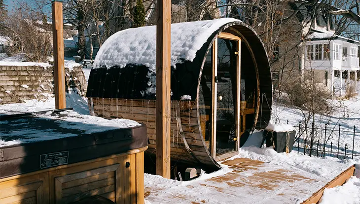 Barrel-shaped outdoor sauna and hot tub surrounded by snow-covered trees.