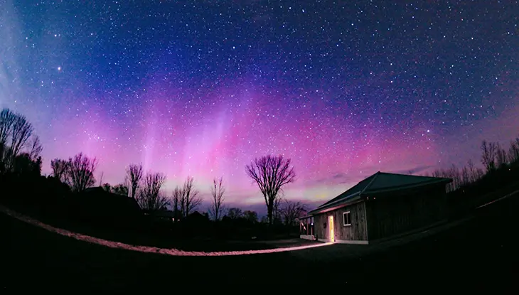 A night sky illuminated by colourful aurora lights above a building and trees.