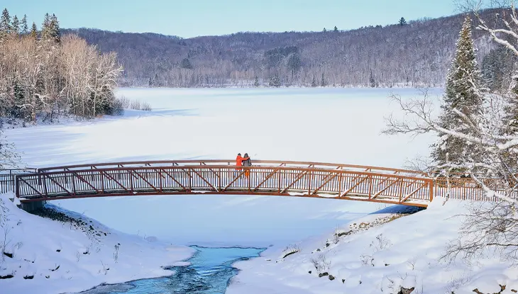 Snow-covered wooden footbridge crossing a partially frozen river surrounded by winter trees.