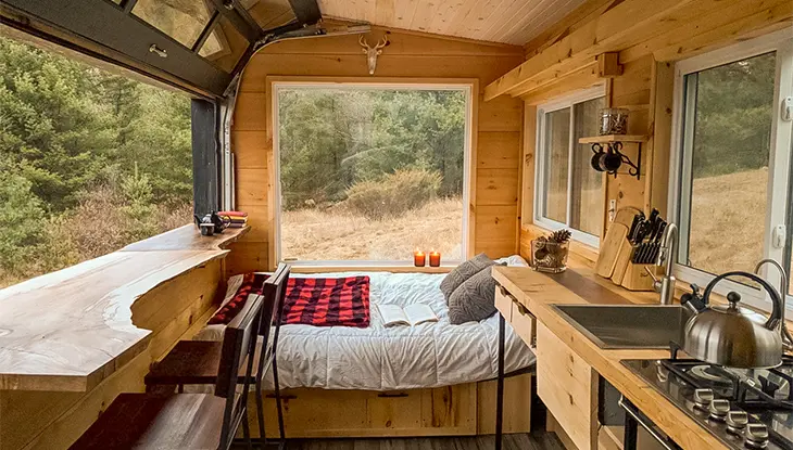 Acogedora habitación tipo cabaña con cama, interior de madera, grandes ventanales y una pequeña cocina con vistas al bosque.