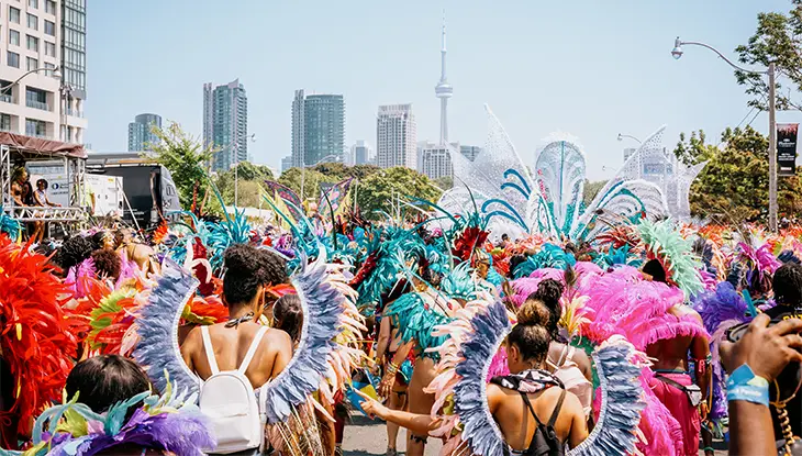 Parade participants wearing feathered costumes march towards downtown Toronto with the CN tower and other large buildings in the distance