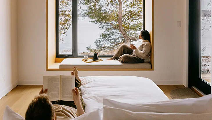 아늑한 침실에서는 호숫가 풍경이 내려다보이며, 한 사람은 침대에서 책을 읽고 있고 다른 한 사람은 물가 옆 큰 창가에 앉아 있다.