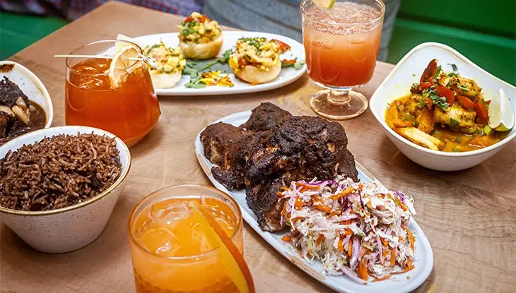 An assortment of Caribbean-style dishes, including rice, grilled meat, and colourful drinks, at a restaurant booth table.