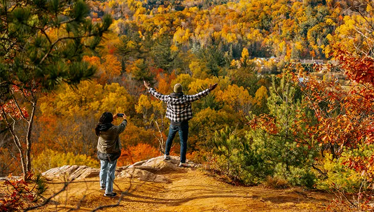 Deux personnes au bord d’une falaise surplombant des arbres aux couleurs d’automne.