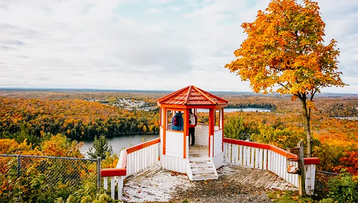 A red and white lookout pavilion on a hilltop overlooking colourful autumn forest and a winding river.