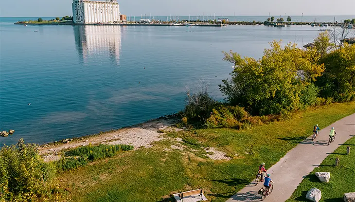 Cyclists ride along a waterfront trail beside the water, with greenery and a large building visible across the water.